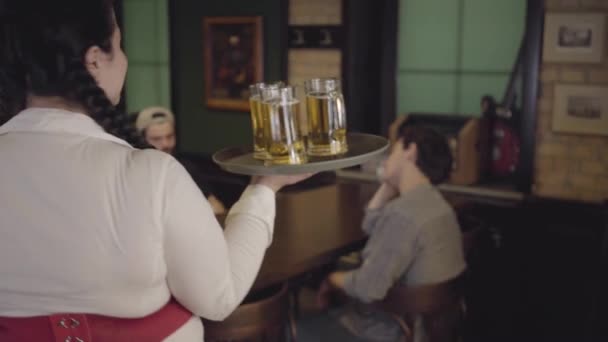 Chica gorda bastante regordeta con tres vasos de cerveza en una bandeja en un restaurante de cerveza. La camarera trae cerveza a dos tipos a los visitantes sentados en una mesa en un bar . — Vídeo de stock