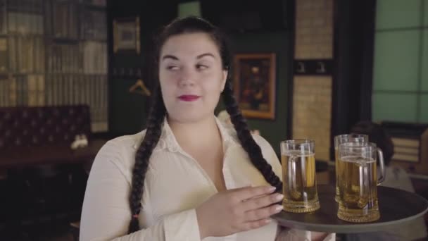 Pulchna kelnerka z warkoczami w białej bluzce zakrada się drinkiem ze szkła klientów. Zabawna dziewczyna nie może się oprzeć i pić pyszne piwo. Wypoczynek w barze — Wideo stockowe
