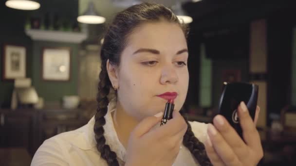 Retrato de atraente mulher gorda confiante pintando lábios com batom vermelho brilhante no restaurante. A menina com duas tranças se preparando para a data, fazendo maquiagem — Vídeo de Stock