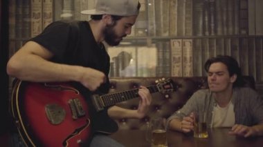 Genç pozitif sakallı adam barda gitar çalarken, onun erkek arkadaşı ritim başını sallayarak yakın oturuyor. Pub 'da boş zaman. Çocuklar sahip eğlenceli birlikte içme bira