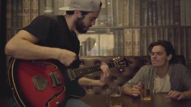 Giovane uomo barbuto positivo che suona la chitarra nel bar, il suo amico maschio seduto vicino scuotendo la testa nel ritmo. Tempo libero al pub. Ragazzi divertirsi insieme bere birra — Video Stock