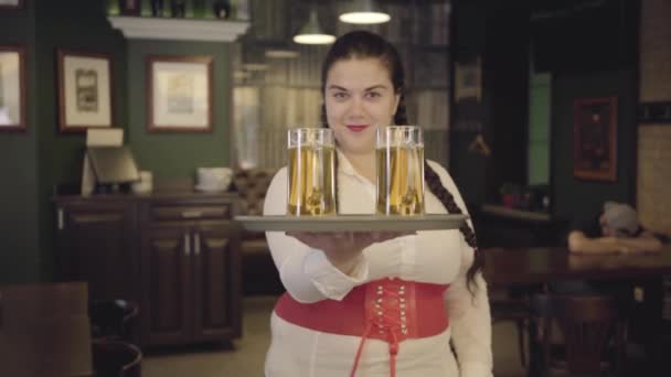 Παχουλός γυναίκα με κοτσίδες σε λευκή μπλούζα κρατώντας δίσκο με δύο ποτήρια μπύρας χαμογελώντας στην κάμερα. Αναψυχή στο μπαρ. — Αρχείο Βίντεο