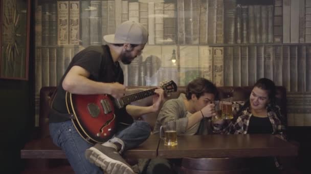 Молодой позитивный бородатый мужчина играет на гитаре в баре, его друзья сидят рядом и пьют пиво. Отдых в пабе. Парни и девчонки веселятся вместе . — стоковое видео