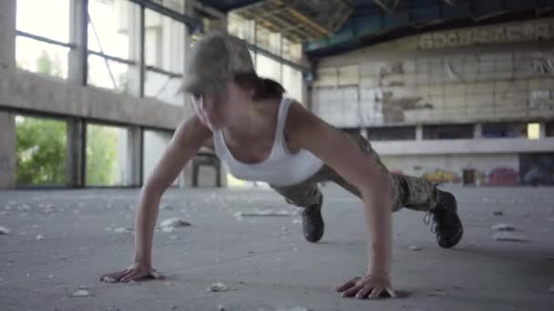 自信的坚强年轻女子穿着军装俯卧撑在一个废弃的工厂里。高跟鞋躺在前景。他的交叉健身锻炼的一部分。高强度间歇训练. — 图库视频影像