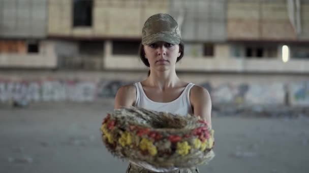 Portrait of a beautiful girl in a camouflage cap and white t-shirt holding a wreath looking at the camera. Warrior woman in a deserted factory. — Stock Video