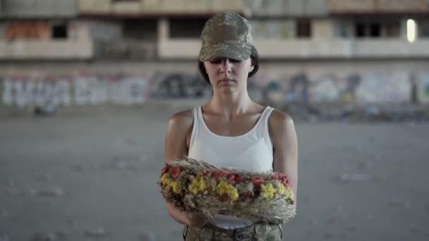 Portrait of a beautiful girl in a camouflage cap and white t-shirt holding a wreath looking at the camera. Warrior woman in a deserted factory. Woman soldier — Stock Video