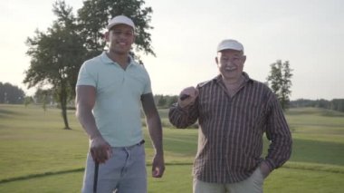 Portre olgun Kafkas adam ve golf alanında Golf oynarken genç Orta Doğu adam. Kamera görünümlü golf oyuncuları gülümsüyor. Yaz tatilinde. Zafer konsepti