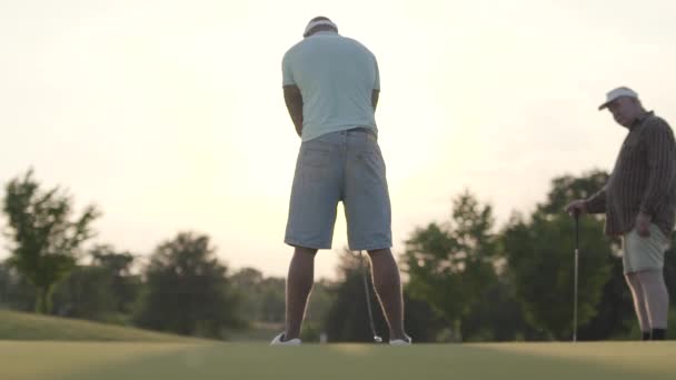 Érett kaukázusi ember és a fiatal közel-keleti ember golfozik a golfpályán. A koncentrált fickó üti a labdát. A koncepció a szabadidő és a sport a szabadban. Victory koncepció. Hátulnézet — Stock videók