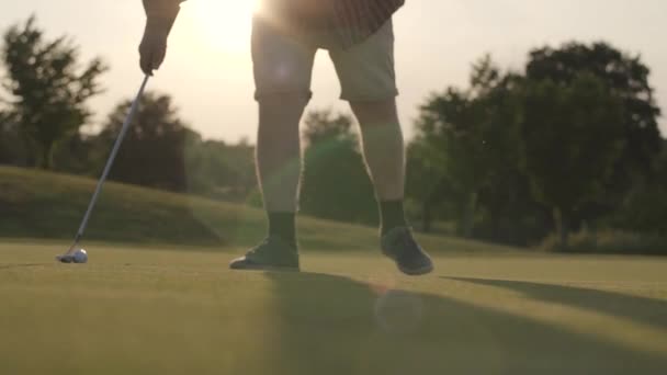 Golfová koule leží na trávě na slunečních paprscích. Nerozpoznaný dospělý muž, který pohybuje míč golfovým klubem. Letní čas. Zpomaleně — Stock video