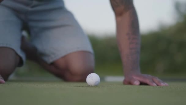 Joven hombre de Oriente Medio jugando al golf en el campo de golf. Hombre guapo soplando en la pelota de golf que se encuentra en el borde del agujero y haciendo que caiga. Ocio de verano — Vídeo de stock