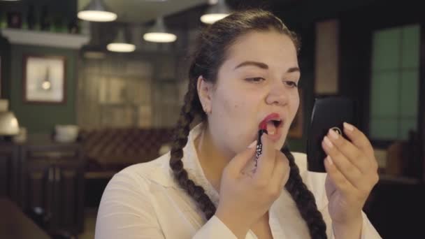 Retrato de atractiva mujer regordeta confiada pintando labios con lápiz labial rojo brillante en el restaurante. La chica con dos coletas preparándose para la cita, haciendo maquillaje — Vídeos de Stock