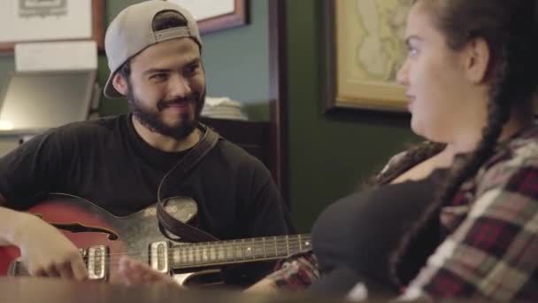 Giovane uomo barbuto sorridente che suona la chitarra nel bar, attraente donna grassoccia seduta vicino a cantare. Tempo libero al pub. Coppia divertirsi insieme — Video Stock