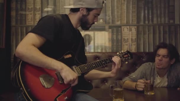 Jonge bebaarde man speelt gitaar in de Bar, zijn vriend zitten in de buurt schudden zijn hoofd in het ritme. Ontspanning in de pub. Jongens hebben plezier samen drinken bier — Stockvideo