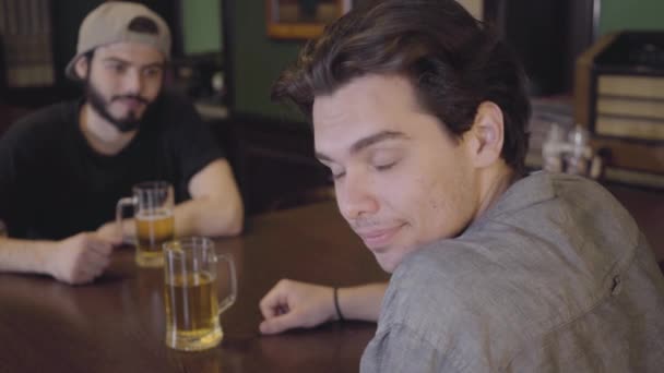 Twee mannen zitten in de bar bier drinken samen. Jongens hebben plezier samen met het drinken van bier. Een man op zoek naar de camera en schudt uitnodigend uitnodigen om mee te doen aan het feest — Stockvideo