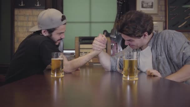 Zwei Männer sitzen an einem Tisch in einer Bar und ringen um den Arm. Der eine gewinnt, der andere verliert, und beide trinken Bier. Jungs haben Spaß beim gemeinsamen Bier trinken — Stockvideo