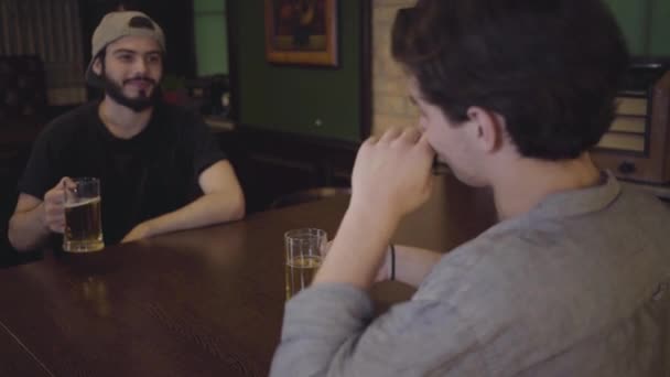 Два парня пьют пиво, сидя за столом в пабе. Парни веселятся вместе, пьют пиво. Отдых в пивном пабе . — стоковое видео