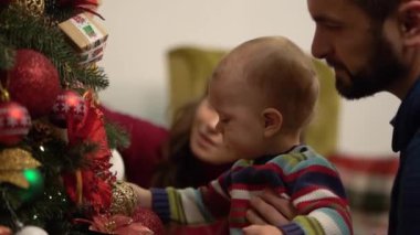 Anne, baba ve küçük bebek dekore Noel ağacı yakın-up yakın oturan. Adam, parlak dekorasyon gösteren köknar ağacında yakın çocuk tutuyor. Mutlu aile Noel 'ı birlikte kutluyor. Yavaş hareket.