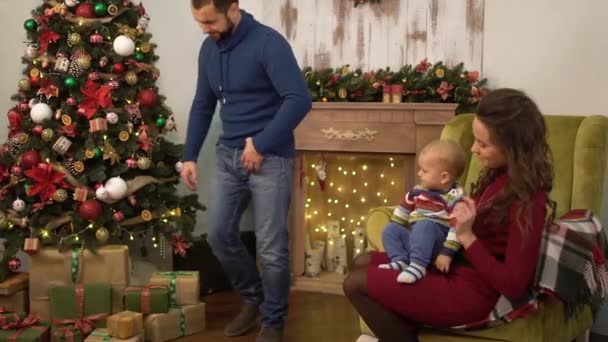 Moeder, vader en kleine baby zitten in de buurt van versierde kerstboom. Man neemt kleine huidige doos en geeft aan het kind zittend op moeder ronden. Gelukkige familie viert Kerstmis samen — Stockvideo