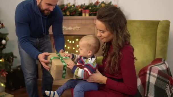 Mother, father and little baby sitting near decorated christmas tree. Dad present box to the child sitting on mother laps. Happy family celebrating Christmas together. — Stock Video