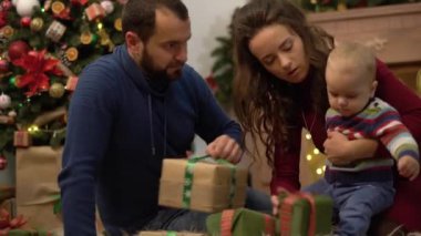 Anne, baba ve küçük bebek Noel dekorasyonu ile odada yerde oturuyor. Adam kabarık halı üzerinde sürünerek çocuk küçük bir hediye kutusu verir. Mutlu aile birlikte Noel kutluyor
