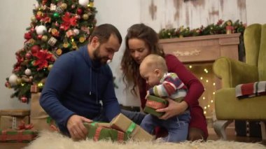 Anne, baba ve küçük bebek Noel dekorasyonu ile odada yerde oturuyor. Çocuk mevcut hediye kutuları ile kabarık halı yatıyor oynuyor. Mutlu aile birlikte Noel kutluyor