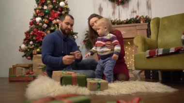 Anne, baba ve küçük bebek Noel dekorasyonu ile odada yerde oturuyor. Babam çocukla oynuyor, oyuncak turuncu dilim elinde tutuyor. Mutlu aile birlikte Noel kutluyor