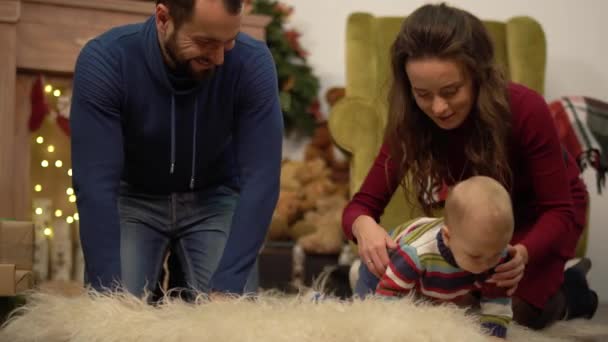 Moeder, vader en kleine baby zittend op de vloer in de kamer met kerst decoratie. Man geeft kleine huidige doos aan kind kruipen op pluizig tapijt. Gelukkige familie viert Kerstmis samen — Stockvideo