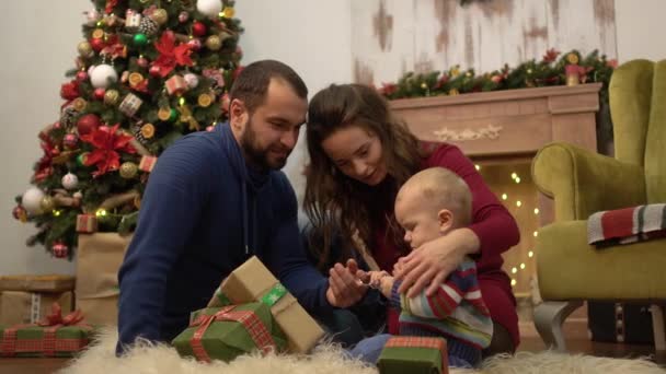 Sevimli eğlenceli aile Noel birlikte anne, baba ve küçük bebek Noel dekorasyonu ile odada yerde oturan kutluyor. Çocuk mevcut hediye kutuları ile oyun kabarık üzerinde yatıyor — Stok video