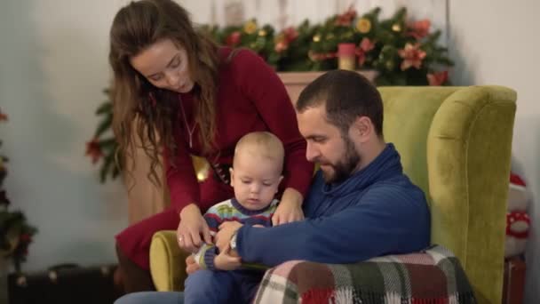 Madre, padre e bambino sono nella stanza con decorazioni natalizie. Uomo che tiene il figlioletto sulle ginocchia, la donna si piega e bacia il marito. Felice famiglia che celebra il Natale insieme — Video Stock