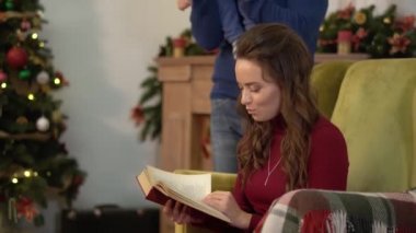 Sevimli büyüleyici genç kadın kocası ve bebek bir sandalye Noel ağacının yanında oturan bir kitap okuma.