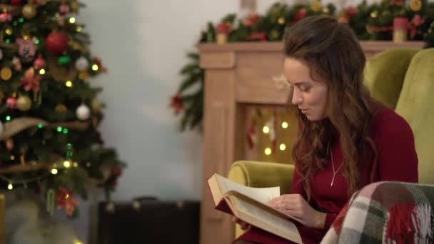 Linda joven encantadora leyendo un libro sentado en una silla junto al árbol de Navidad . — Vídeos de Stock