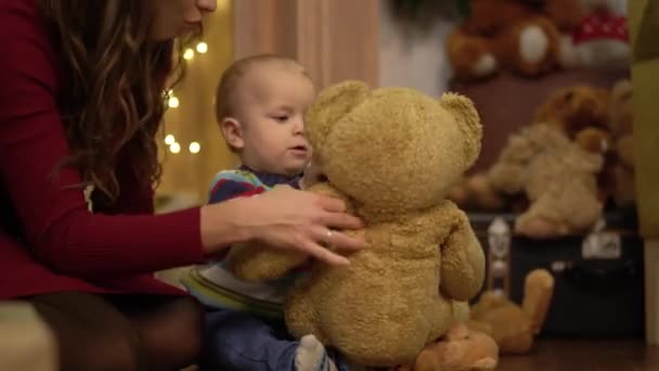 Close up view of little cute child playing with big teddy bear at home. Mother sitting near the son holding shaking toy and helping her baby. Concept of parenthood, motherhood — Stock Video