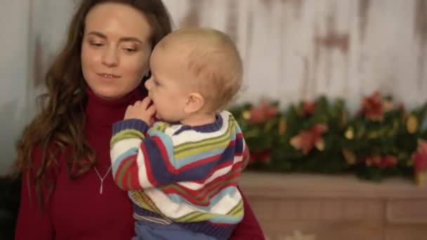 Mujer joven y bonita sacude al bebé en sus brazos de pie cerca del árbol de Navidad y muestra juguetes brillantes decoraciones para el hijo. Feliz familia celebra la Navidad — Vídeos de Stock