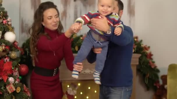 Mãe, pai e um bebê se divertem na sala com decoração de Natal. O pai e a mãe brincam com a criança, atira-o de braços erguidos. Família feliz celebrando o Natal juntos — Vídeo de Stock