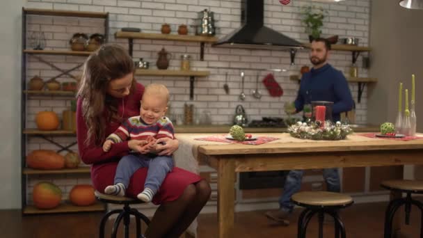 Young pretty woman sitting at the table in the kitchen with baby on her pals while handsome bearded man bringing her glass of lemonade drink. Happy family spend time together — Stock Video