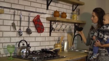 Modern mutfakta kollarında bir bebek ile güzel genç kadın kahvaltı hazırlanıyor. Lady yemek karıştırmak için kase ve büyük kaşık alır. Mutlu aile konsepti