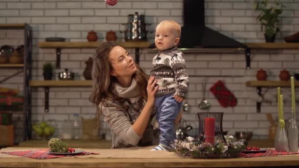 Jolie jeune femme soulève le bébé dans ses bras debout dans la cuisine moderne montrant au fils jouet rouge vif arbre de Noël suspendu au plafond. Concept de famille heureuse — Video