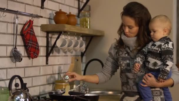 Pretty young woman with a baby in her arms in modern kitchen preparing breakfast. Lady pours salt into the frying pan cooking food. Happy family concept — Stock Video