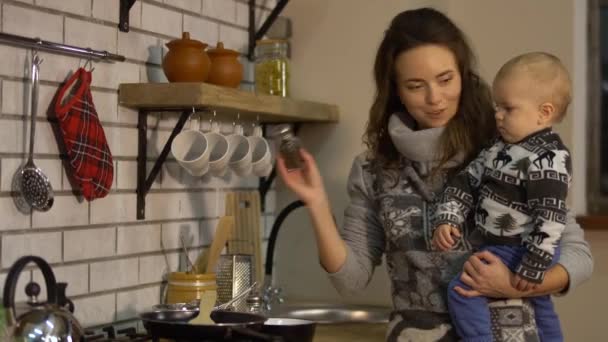 Jolie jeune femme avec un bébé dans les bras dans la cuisine moderne préparant le petit déjeuner. Dame verse du sel dans la poêle à frire cuisson des aliments. Concept de famille heureuse — Video