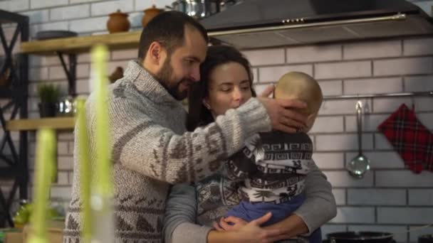 Pai barbudo em camisola quente brincando com o filhinho que está nos braços da mãe na cozinha. A mulher dá o filho ao pai e leva-o de volta. Feliz família amigável passar tempo juntos — Vídeo de Stock