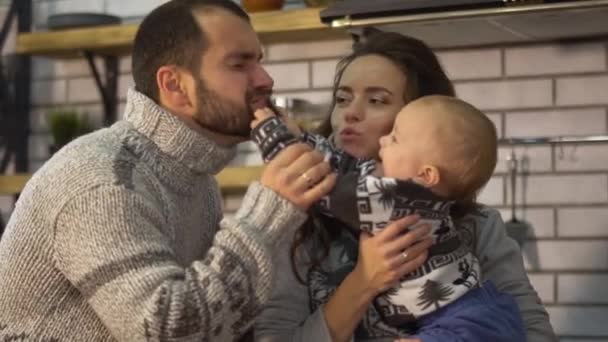 Smiling father in warm sweater playing with baby little son who is in mother arms in the kitchen. Child tugging dad by the beard. Happy friendly family spend time together — Stock Video