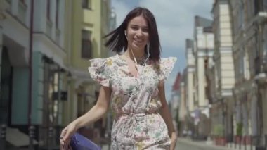 Alışveriş çantaları ile sevimli neşeli genç kadın. Şehir Sokağı boyunca dans ederken kulaklık ve cep telefonu ile kız müzik dinler. Genç eğlenceli kız açık havada zevk. Yavaş hareket.