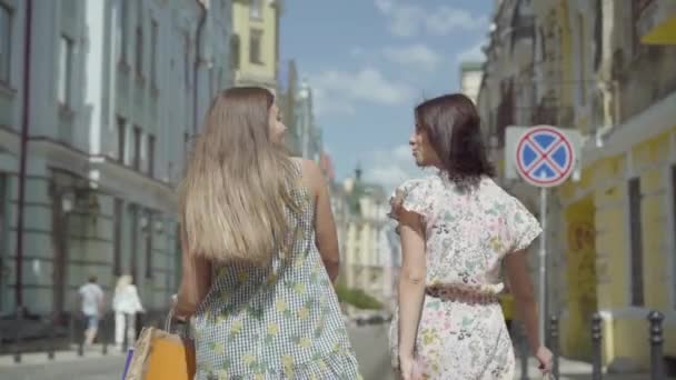 Twee vrolijke vrouwen met boodschappentassen wandelen door de stads straat. Jonge meisjes dragen stijlvolle zomer jurken genieten met tijd doorbrengen. Shopping lifestyle concept. Slow Motion. — Stockvideo