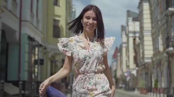 Mulher jovem alegre bonito com sacos de compras. Menina com fones de ouvido e telefone celular escuta música enquanto dança pela rua da cidade. Jovem menina divertida desfrutando ao ar livre. Movimento lento . — Vídeo de Stock