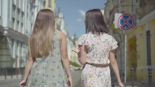 Achteraanzicht van twee wandel vrouwen met boodschappentassen. Jonge meisjes dragen stijlvolle zomer jurken genieten met tijd doorbrengen. Shopping lifestyle concept. Slow Motion. — Stockvideo