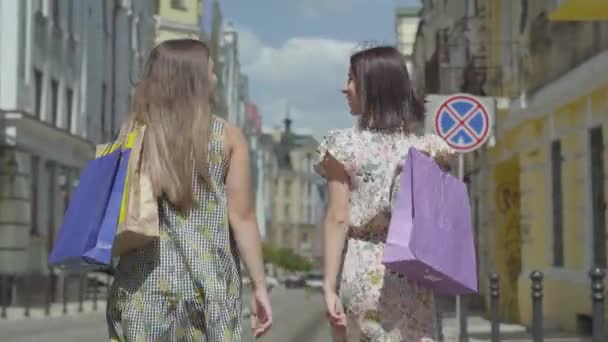 Achteraanzicht van twee wandel vrouwen met boodschappentassen. Jonge meisjes dragen stijlvolle zomer jurken genieten met tijd doorbrengen. Shopping lifestyle concept. — Stockvideo