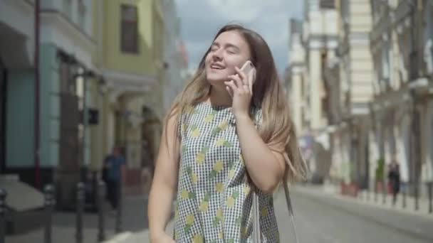 Adorável jovem mulher falando por telefone celular andando pela rua da cidade. Menina divertida vestindo vestido de verão elegante desfrutando ao ar livre. Movimento lento . — Vídeo de Stock