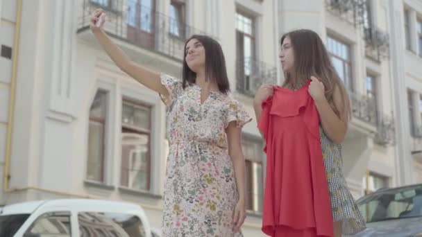 Zwei süße Freundinnen nach dem Einkaufen mit Einkaufstaschen Selfie auf dem Handy mit neuem Kleid im Freien. Freizeit glücklicher Mädchen. Unbekümmerte Damen spazieren durch Stadtstraße. — Stockvideo