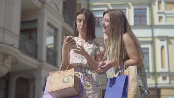 Две очаровательные счастливые подружки после покупок с сумками для покупок, переписывающимися по телефону перед красивыми зданиями. Отдых oung девушки носить стильные летние платья наслаждаясь проводить время — стоковое видео