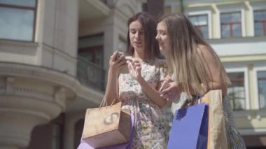 İki sevimli kız alışveriş torbalar ile alışverişten sonra telefon açık havada selfie alıyor. Mutlu kızların boş olması. Şehir sokakta yürürken Carefree bayanlar.